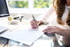 a woman signs a document while sitting at a table using a laptop